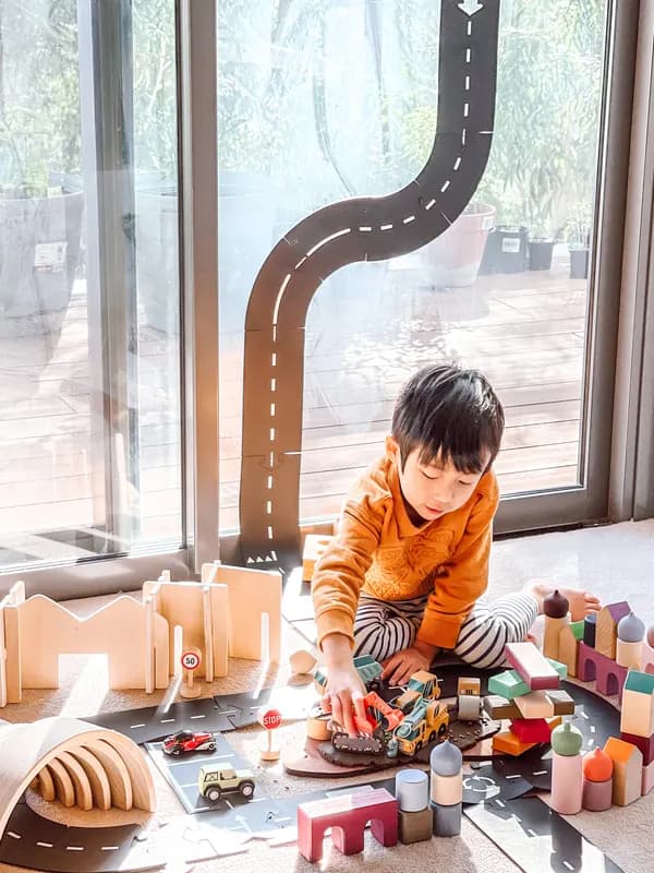 A boy playing with a wooden road set and toy cars by the window