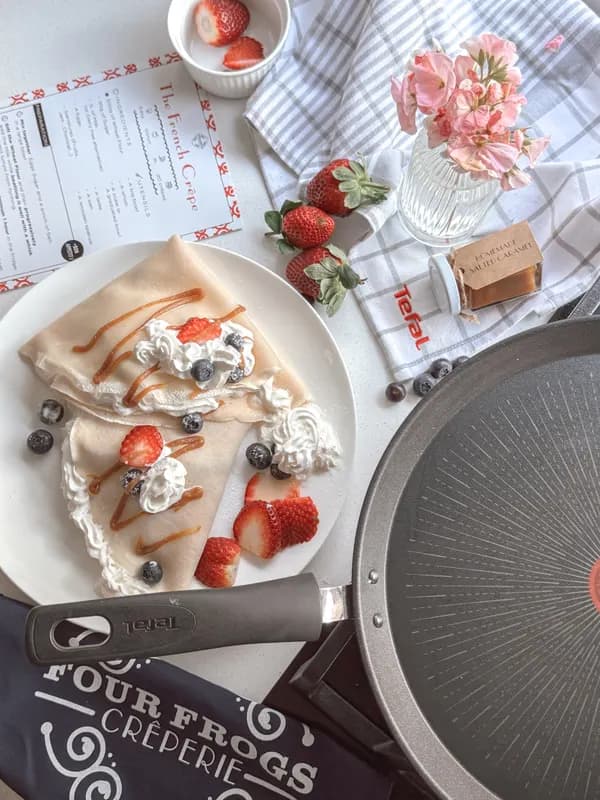 A flat lay of crêpes with strawberries and cream beside a pan
